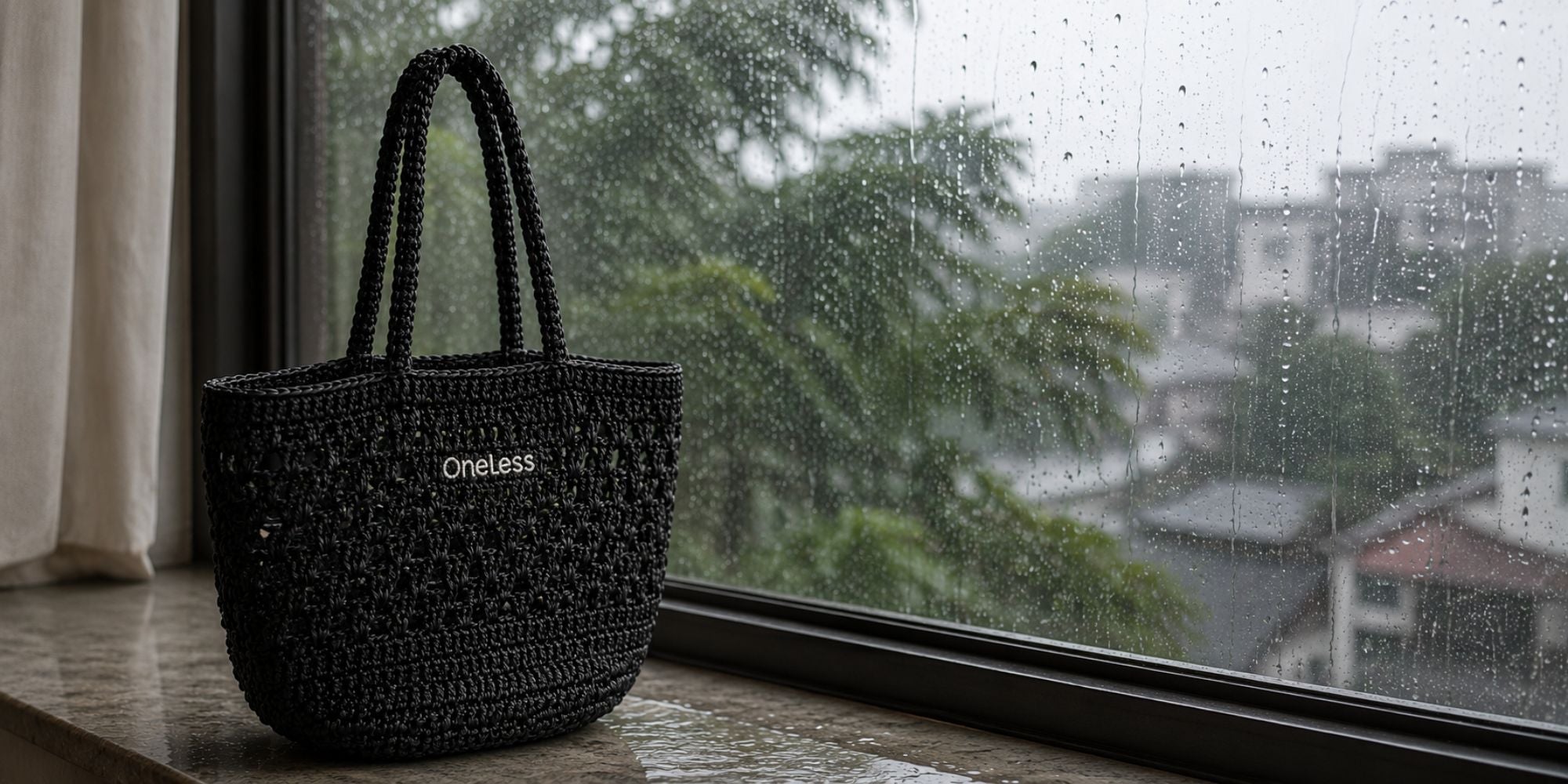 One Less black woven raffia tote against a rainy window, representing monsoon moisture and bag care